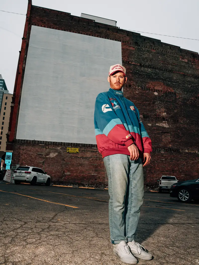 Zewmob standing in a parking lot beside a brick wall in a vintage racing jacket and light-wash jeans